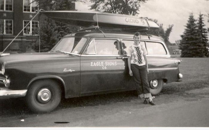 A black and white photo of a woman standing next to a car with a canoe on top.