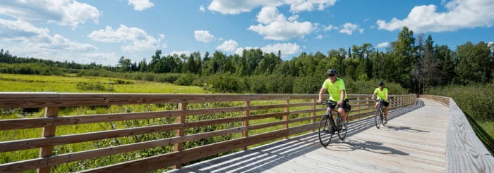 Two people riding bikes on a wooden boardwalk in a grassy area.