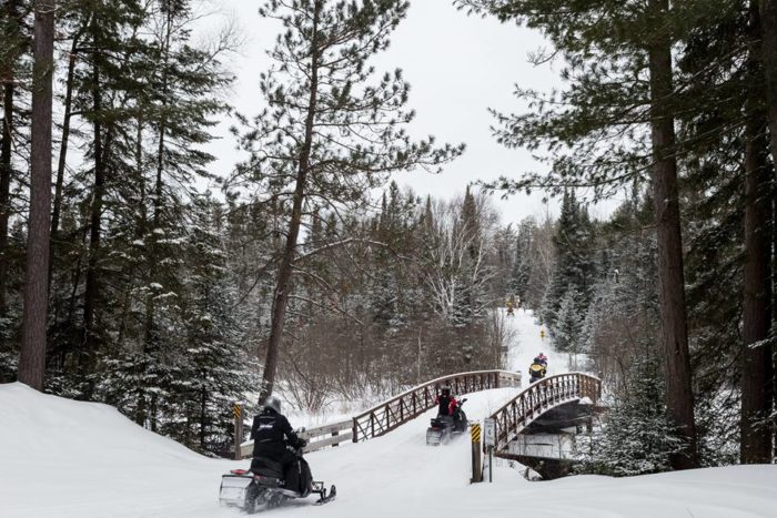 A group of people riding snowmobiles on a bridge in the woods.