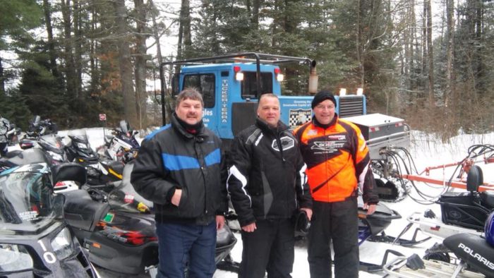 Three men standing next to snowmobiles in the snow.