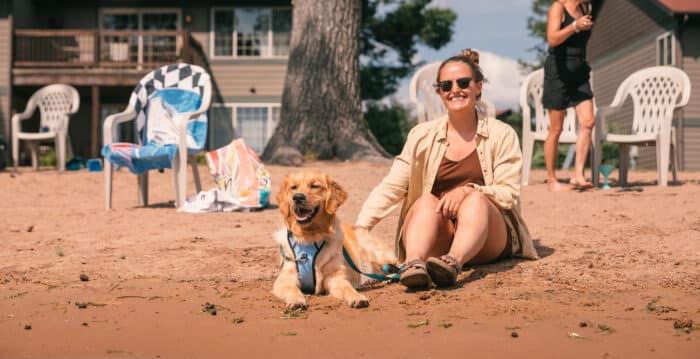 A woman sits on the sand next to a golden retriever wearing a harness, with empty chairs and lakeside houses in the background.