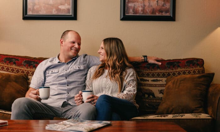 A man and woman sit on a patterned sofa holding mugs, smiling at each other, with framed artwork on the wall behind them.