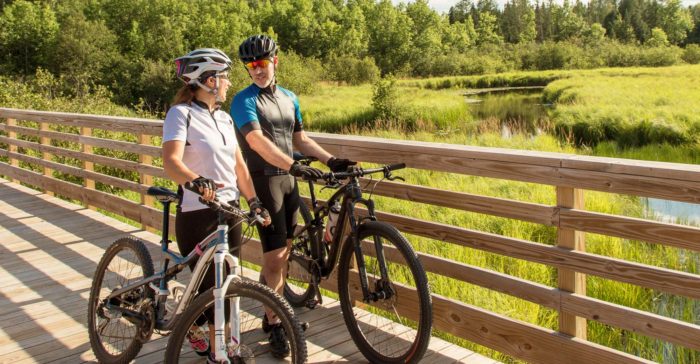 Two bicyclists standing on a wooden bridge near a pond.