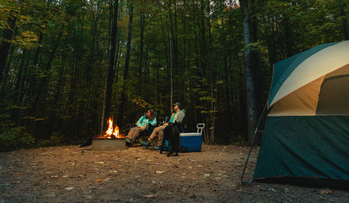 Two people sitting around a campfire in the woods.