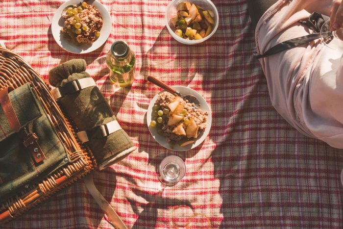 A woman sits on a picnic blanket with a basket full of food.