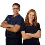 Two individuals in medical scrubs, a man and a woman, stand side by side, both with arms crossed and smiling. The man wears glasses. The background is plain white.