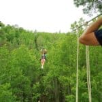 A person wearing a harness rides a zip line over a forested area, with trees and greenery visible below and around them.