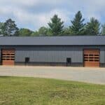 A long, black and brown metal building with four large garage doors stands on a gravel lot bordered by grass, set against a backdrop of trees and a cloudy sky.