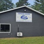 A single-story building, framed by trees, displays a sign that reads "ASAP Garage Doors" along with contact numbers 715-545-3172 and 715-479-2728.