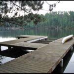 A wooden dock, crafted from fine wood products, extends over a calm lake. A canoe is upside down on the right side of the dock. Trees line the opposite shore under a cloudy sky.