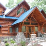 A large log cabin with a blue metal roof features stone accents and a covered porch. Surrounded by trees and greenery, this charming retreat showcases the finest wood products for a rustic yet refined ambiance.