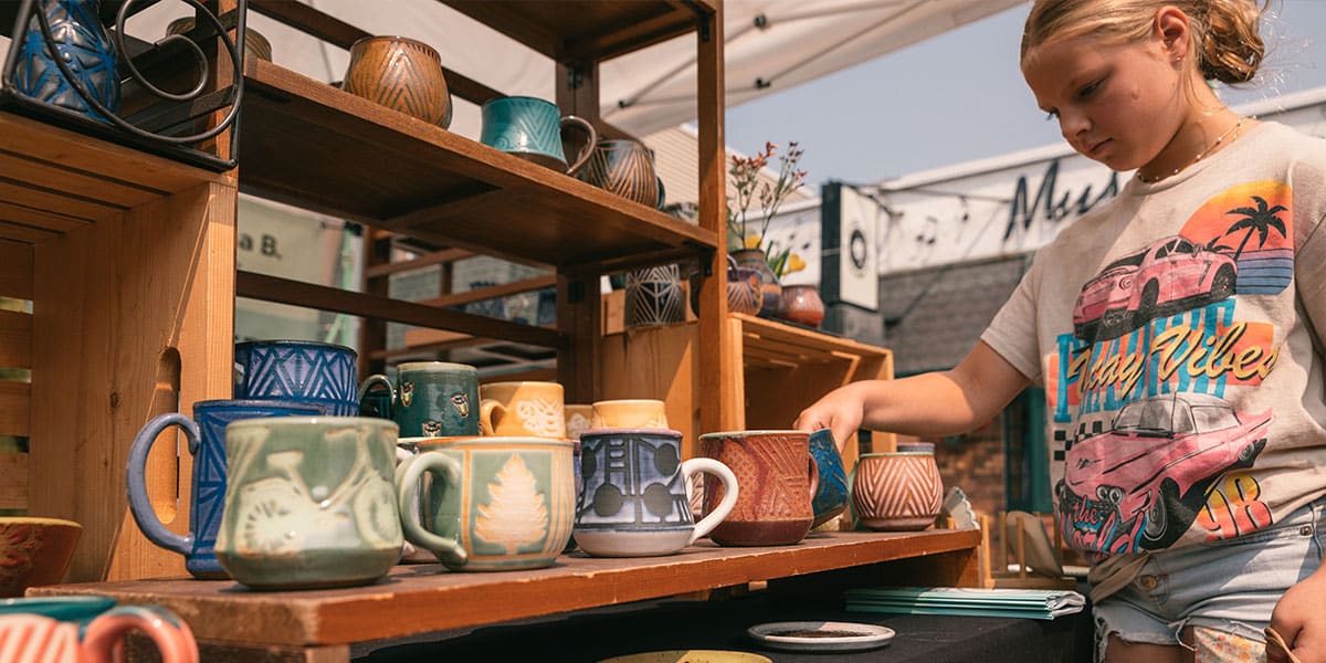 A young girl examines colorful mugs displayed on wooden shelves at an outdoor market.