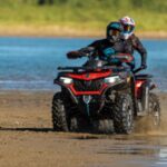 Two people wearing helmets ride an ATV rental on wet sand near a body of water, with mud and water splashing behind them.