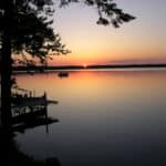 A tranquil lake at sunset with a boat near a dock at the resort, surrounded by silhouettes of trees. The sky displays a gradient of colors from orange to purple.