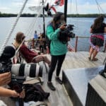 People are filming and recording a scene on a boat. Two cameras are focused on a woman sitting on a deck, while another person adjusts an audio device. A large body of water and trees are in the background.