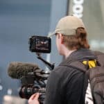 Person wearing a cap and backpack operating a professional video camera with an attached microphone and monitor.