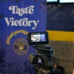 A video camera on a tripod records a scene in front of a blue and yellow "Taste Victory" banner with a logo.