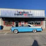 A blue and white classic convertible car is parked in front of the Warehouse Art Center building on a sunny day.