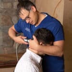 A chiropractor in blue scrubs uses an adjusting instrument on a seated man's neck in a professional office setting with stone wall decor.