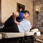 A chiropractor in a blue shirt applies pressure on the back of a patient lying on their side on a treatment table in a rustic office.