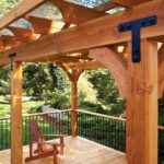 A wooden deck featuring a stylish pergola and a single wooden chair, all crafted from high-quality wood products, overlooks a lush garden area with trees and bushes in the background.