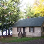 A small beige house with a green roof, surrounded by trees, exudes the charm of a secluded resort. There are stairs leading to the front door, and a lake is visible in the background.