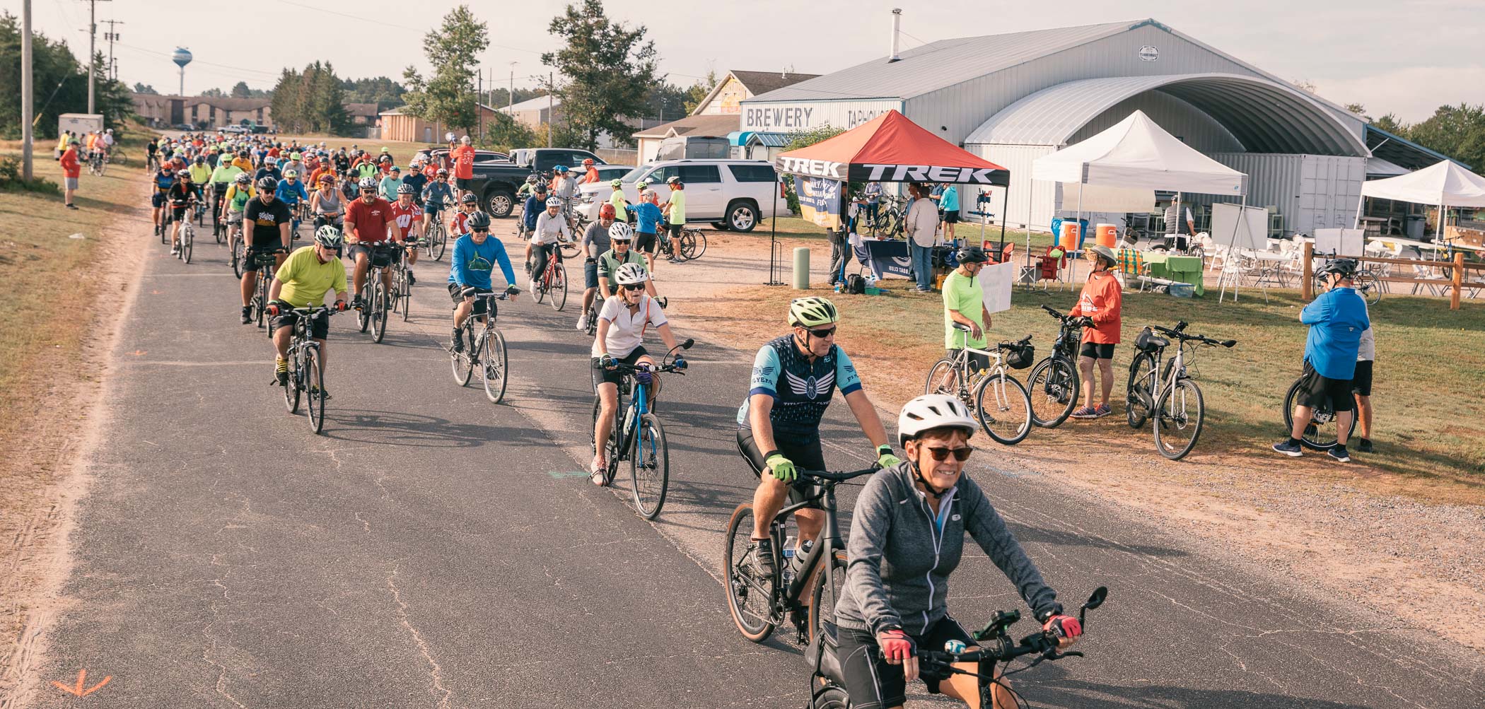 A group of cyclists participates in a biking event on a country road, passing by a brewery and a Trek tent.