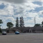 A nearly empty parking lot of a single-story building offers a serene environment, with just a few cars parked. The sky is cloudy, and trees surround the area. On the left, a signboard for physical therapy stands prominently amidst this calm setting.