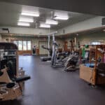 A gym with exercise equipment including treadmills, weight machines, and elliptical trainers designed for workouts and physical therapy. A few chairs are placed along the wall.