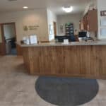 Reception area with a wooden desk, computers, and information displays. A hallway leads to additional rooms dedicated to physical therapy. Walls are decorated with a logo and various documents.