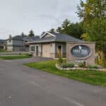 A paved driveway leads to residential buildings with attached garages at Eagle Trail Estates, a prime property rental. A stone sign displays the property's name and contact number, surrounded by landscaped greenery.