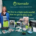 A woman stands beside a table displaying brochures and promotional items for Norvado, a company offering internet, television, telephone, and managed services, in front of a branded green backdrop.