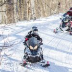 Two people ride snowmobiles on a snowy trail through a forest of leafless trees, enjoying an exhilarating powersport rental adventure.
