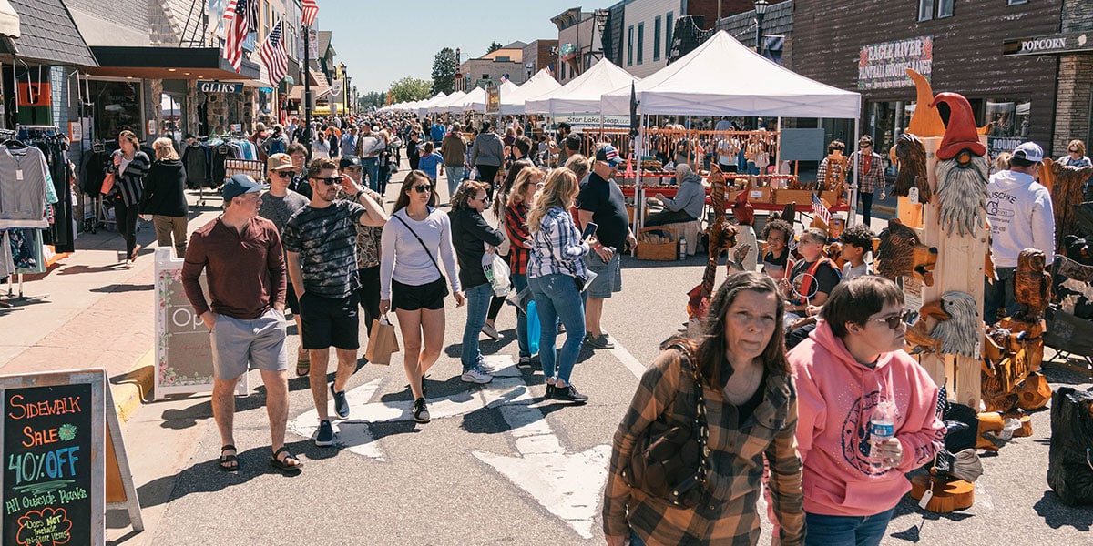 A busy outdoor market on a sunny day with people strolling past vendor tents and a variety of wooden carvings on display.