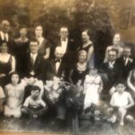 A sepia-tone photograph of a large multi-generational family, some seated and some standing, posed outdoors with a backdrop of trees. Promotional products are nestled among two large floral arrangements at the center front.