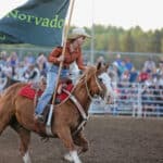 A rodeo participant on horseback carries a flag that reads "Norvado" in front of a bleacher full of spectators.