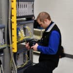 A technician kneels while working on network equipment in a server room, connecting cables to a rack-mounted switch.