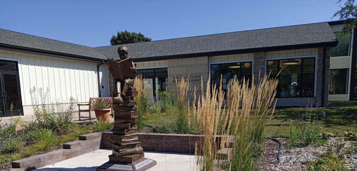 A bronze statue of a child reading a book stands in a garden outside the St. Olaf Library, capturing the spirit of learning on a sunny day.