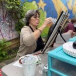 An older woman with gray hair sits at a table outdoors, painting on a canvas. She wears glasses and a beige sweater. A colorful mural is on the wall in the background—part of the vibrant display from the nearby Art Center. Art supplies are scattered across the table, each adding to her creative process.