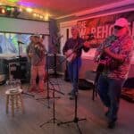 Four musicians perform on a small stage at the Art Center with a banner reading "THE WAREHOUSE" in the background. They are playing a banjo, guitar, fiddle, and bass under colorful lights. A fishbowl rests on a stool in front.