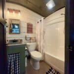 A small bathroom with a sink, toilet, and shower-tub combo, decorated with rustic accents, patterned curtains, and a wall sign above the toilet creates a cozy resort-inspired retreat.