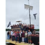 A group of people stands on a dock in front of a pirate-themed boat with flags and a mast on a cloudy day, discussing accounting strategies for their next adventure.