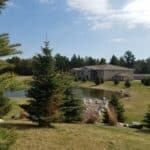 A two-story house with beige siding is available for property rental near a small pond, surrounded by green lawns, trees, and rocks under a partly cloudy sky.