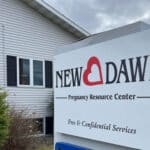 A sign for New Dawn Pregnancy Resource Center stands outside a light grey building with a bush to the left. The sign highlights free and confidential pregnancy resource services.