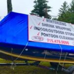 A blue boat covered with a blue tarp sits on a trailer; a large sign advertises winterizing, shrink wrapping, storage, cleaning services, and boat rental with a phone number.