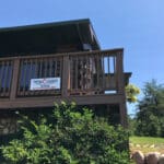 A sign labeled "New Dawn Office" is affixed to the balcony railing on a wooden building. Below, there is a stone foundation partially covered by greenery, hinting at the supportive environment of this pregnancy resource center. The sky above is clear and blue.