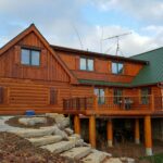 A large, two-story wooden cabin crafted from premium wood products with a green metal roof, elevated on tall pillars with a spacious deck, sits in a rustic outdoor setting. Stone steps lead to the entrance. Trees surround the area.