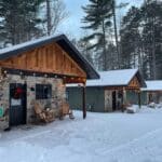 Three small stone and wood resort cabins with snow-covered roofs sit in a snowy forest area. Wooden chairs and seasonal decorations adorn the porches, creating a cozy winter retreat.