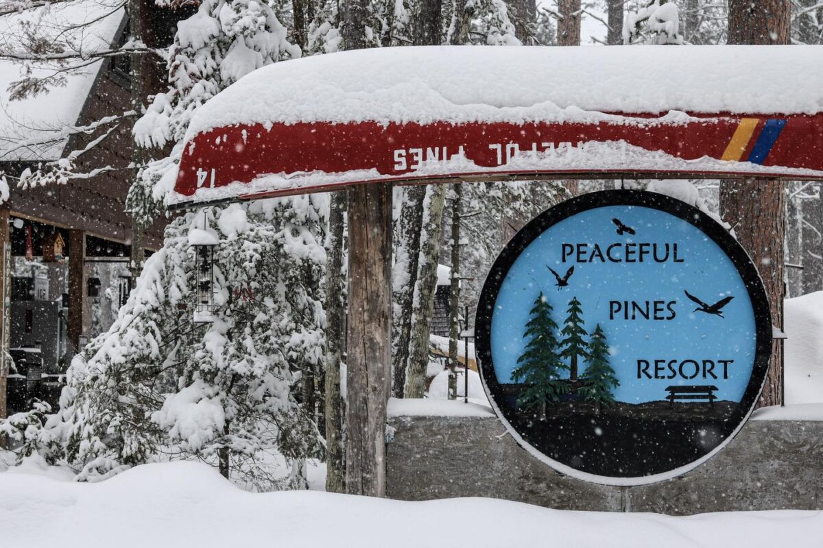 A snow-covered canoe rests above a sign reading "Peaceful Pines Resort," surrounded by snowy trees and the inviting glow of a cozy resort cabin in the background.
