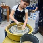 A young girl in an art center's pottery studio shapes clay on a potter's wheel, with her hands covered in wet clay. Another person stands nearby, with pottery supplies and materials in the background.